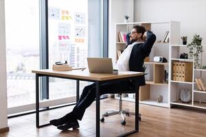 Handsome adult male office worker wearing suit sitting relaxed in professional workspace, bright modern office with city view background, enjoying break from work and contemplating ideas. photo