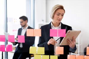 Businesswoman and businessman in professional attire working with sticky notes and devices indoors. Scene represents teamwork, planning, modern business environment, creativity, and productivity. photo