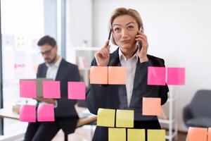 Professional businesswoman multitasking with phones and managing tasks using colorful sticky notes on transparent board. In the office setting, male colleague collaborating in background. photo