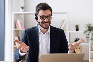 Smiling young man in formal suit using headset and laptop during call in office. Professional working environment with bookshelves in background showcases cheerful approach to remote working. photo