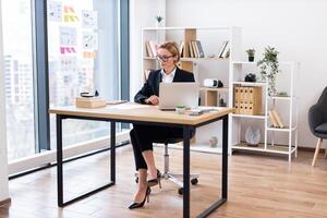 Middle-aged Caucasian woman working on laptop in bright modern office setting, symbolizing productivity and workstation organization photo