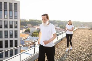 Senior male and female jogging on city rooftop. Active lifestyle, health, and wellness among urban seniors promoting fitness and well-being. Sunlight enhances cityscape and adds warmth to scene. photo