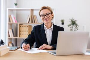 Professional woman wearing business attire writing on paper at desk with laptop in office. Smiling face suggests positivity, productivity. Background shelves hold books plants and organized documents. photo