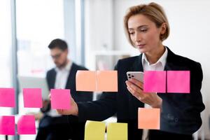 Professional adult caucasian businesswoman interacting with colorful sticky notes on glass board while man working in background in modern office environment photo