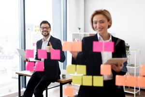 Man and woman in business attire collaborating on project using sticky notes on glass board. Modern office setting with teamwork and productivity focus. photo
