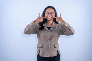 A young woman in a brown jacket and glasses is showing a wide smile with fingers pointing at her cheeks happily. photo