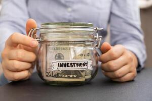 Unrecognizable woman holding Saving Money In Glass Jar filled with Dollars banknotes. INVESTMENT transcription in front of jar. Managing personal finances extra income for future insecurity background photo