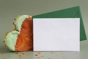 Empty template paper note with envelope greeting card mock up. Heart shaped pistachio croissant on green background with copy space. Puff pastry new trendy sweet dessert bakery. Creamy dough experiment photo