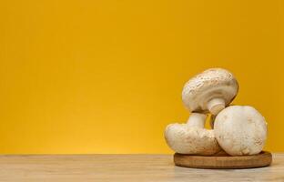 Fresh champignons with white caps and light brown stems, arranged on a round wooden stand on a table. The background is bright yellow photo