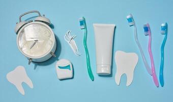Set of items for oral hygiene, white tube of toothpaste, toothbrushes, dental floss and white alarm clock, and decorative paper teeth, on blue background. Top view. photo