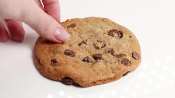 A hand reaches for and picks up a chocolate chip cookie from a white surface under bright lighting video