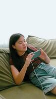 Asian girl lying on green sofa using smartphone with headphones, focused expression home interior with soft light from window technology gadgets youth lifestyle, vertical video