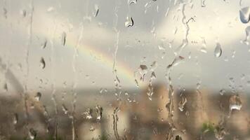Raindrops running down the window during the daytime rain against the backdrop of rainbows and gray clouds over the city video
