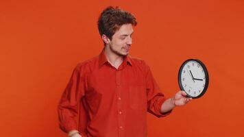 It is your time, hurry up. Caucasian man showing time on wall office clock, ok, thumb up, approve, pointing finger at camera, advertisement, deadline. Young guy isolated on studio orange background video