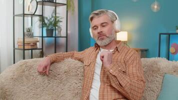 Caucasian mature man sitting on sofa at home, wearing wireless headphones listening to an audiobook. Middle-aged guy is studying language through online learning, appearing immersed in story, enjoying video