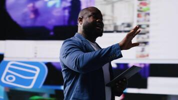 Black presenter hosting conference on stage surrounded by media visuals, symbolizing global convention with live audience to discuss about online trends and mobile apps. Public event. Camera B. video