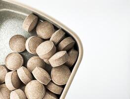 Brown small round pills lie in a silver rectangular pillbox on a white background on the left in the image top view photo