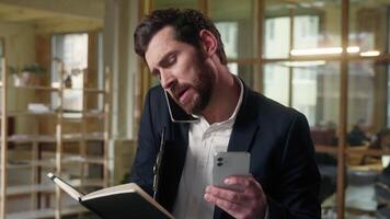 Multitasking businessman guy business man talking mobile phone in office use gadgets consultation Caucasian male employer busy entrepreneur broker planner organizer with notebook manager working call video