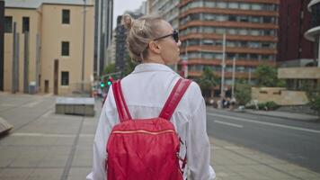 A woman with a red backpack walks along a city sidewalk, surrounded by modern buildings and urban traffic. She is dressed in a white shirt and a floral skirt, and she holds a phone in her hand. video