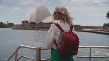 A female tourist with a red backpack stands at the edge of a city ferry, with the iconic Sydney Opera House visible in the background. Showcases harbor public transport with world-famous landmarks. video