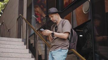 A middle-aged man in casual attire with a backpack stands on urban stairs, checking his smartphone for transport apps and maps. Captures modern city navigation video
