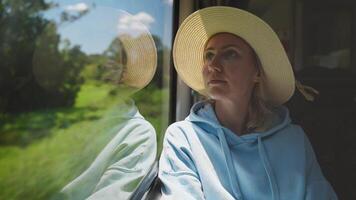 A woman wearing a straw hat and a light blue hoodie sits by the train window, gazing outside with a thoughtful expression. video
