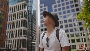 A man wearing a black hat, white t-shirt, and a backpack is standing in the middle of a modern city, looking up at the tall glass and concrete buildings surrounding him. video