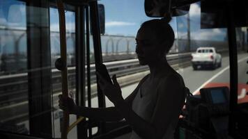 A dark silhouette of a young woman stands in a city bus, holding a smartphone while illuminated by passing highway lights. Through the window, a busy urban freeway pulses with nighttime traffic video