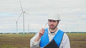Portrait of young and talented engineer, checking the work at wind farm. He is wearing hard hat and glasses, and has folder with documents in his hand. video