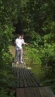 A young couple stands on a wooden village bridge in the forest, holding hands and looking at the river. They hug and have a tender moment. video