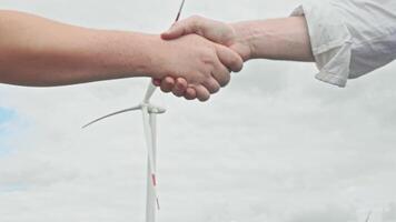 Two partners shake hands after signing contract against background of sky and wind turbine that rotates and generates clean, environmentally friendly energy. video