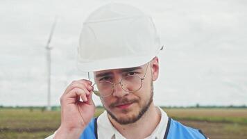 Portrait of a professional engineer, wearing glasses, preparing to start inspecting wind turbines, standing in the middle of an agricultural field. headshot video