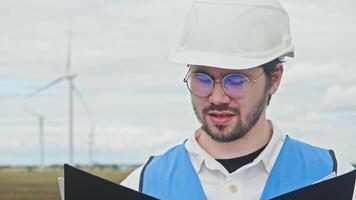 Chief engineer, wearing a construction helmet, glasses, and vest, studies the documentation and drawings for a wind farm located in an agricultural field. Portrait. video