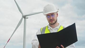 Young engineer in a vest and helmet is admiring an environmentally friendly wind turbine in the field. He gives a thumbs-up and smiles at the camera. video
