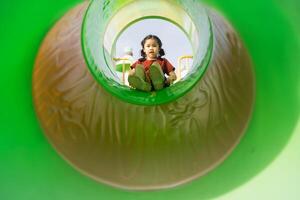 Joyful Child Playing in Colorful Playground Slide Captured from Inside Viewpoint with Bright Green Surroundings, Fun Outdoor Activity Daytime photo