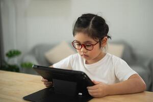Young girl wearing glasses, focused on tablet screen while sitting at a wooden table in a bright, modern indoor setting with soft furnishings photo