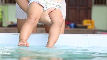 Mother gently holding her baby, dipping the tiny feet into water. Warm and tender moment, symbolizing love of mom, care, bonding, early sensory experience, and playful connection with nature, close-up video