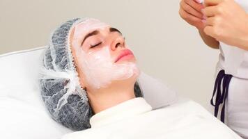 A professional cosmetologist makes a fabric moisturizing mask on the face in a beauty salon on a light background. The concept of spa and relaxation. photo