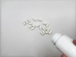A man poured out white pills for illness lying on a white background spilled out of a white medicine jar onto a flat white surface the jar is located at the bottom right in the image view from above photo