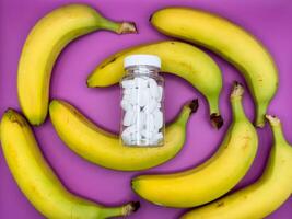 Background of bright yellow bananas and white pills in a transparent jar on a purple background, top view photo