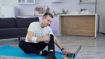 Young man sitting on a mat after training in front of a laptop. Sportive guy talking to someone through the laptop and shows a bottle of water in the kitchen. video