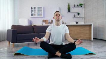 Young man sitting in yoga pose in the kitchen. Guy in white t-shirt doing relax exercises after workout while sitting on a mat at home. video