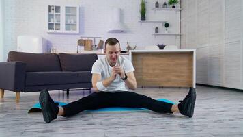 Young man doing workout at home. Guy is sitting on a mat and trying to do exercising during a quarantine period. Difficulties of doing workout at home. video