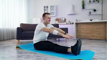 Young man exercising at home. Man sitting on a mat and trying to touch his feet on kitchen background. Difficulties of physical training during lockdown period. video