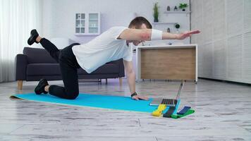 Guy in white t-shirt and black pants training in the kitchen. Young man doing exercise on a mat and looking into a laptop. Slow motion. video
