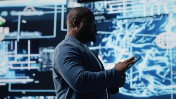 Black male IT engineer examining digital AI brain interface on dashboard, symbolizing neural networks, science and algorithmic thinking. Cybernetics for high tech machine learning. Camera B. video