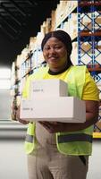 Vertical Black woman managing the order fulfillment process by checking packages in a large business depot filled with racks. Employee oversees operations for retail e-commerce express delivery. Camera A. video