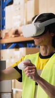 Vertical Worker uses virtual reality headset to ensure order fulfillment with AI, tracking parcel status for e-commerce logistics. Asian man in industrial depot with racks and boxes. Camera B. video