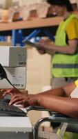 Vertical African american wheelchair user in safety vest processing cargo orders on pc at desk, logistics center optimized for reduced mobility. Female clerk handles online shopping operations. Camera B. video