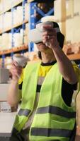 Vertical Asian man uses virtual reality headset and tracking software near industrial storage racks, processing orders. VR device to update tracking info on parcels, artificial intelligence. Camera A. video